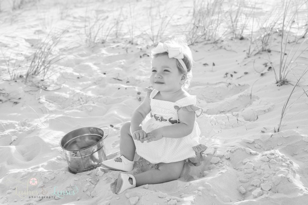 A black and white photo of a one year old girl sitting in the sand with a bucket at Johnsons Beach in Perdido Key