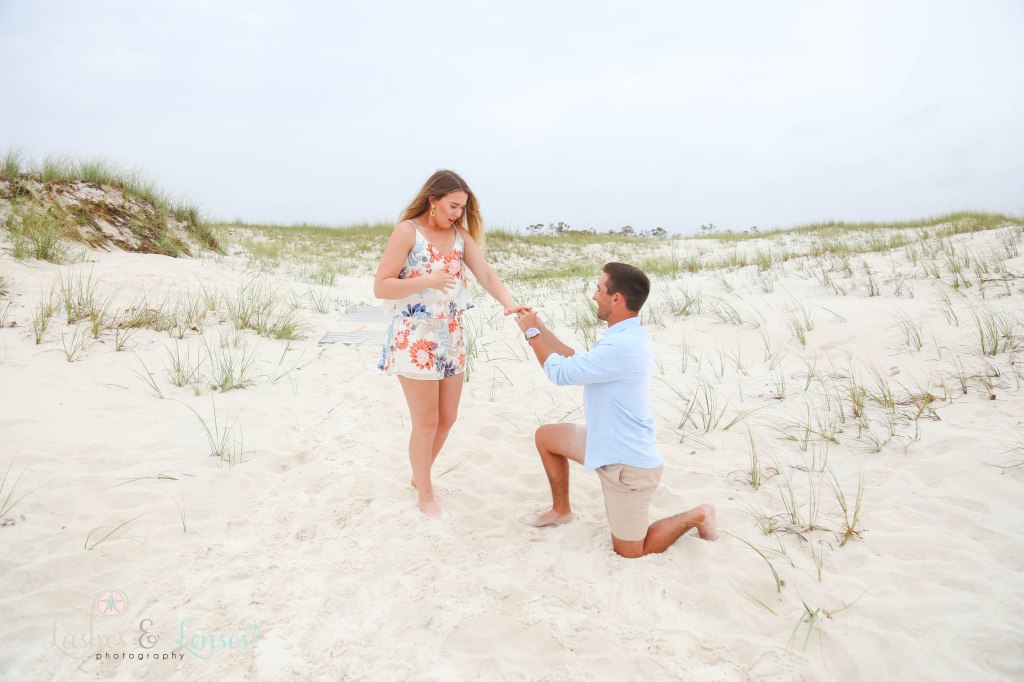 Surprise Engagement Session, man putting engagement ring on girlfriends hand with the sand dunes behind them at Johnsons Beach in Perdido Key Florida