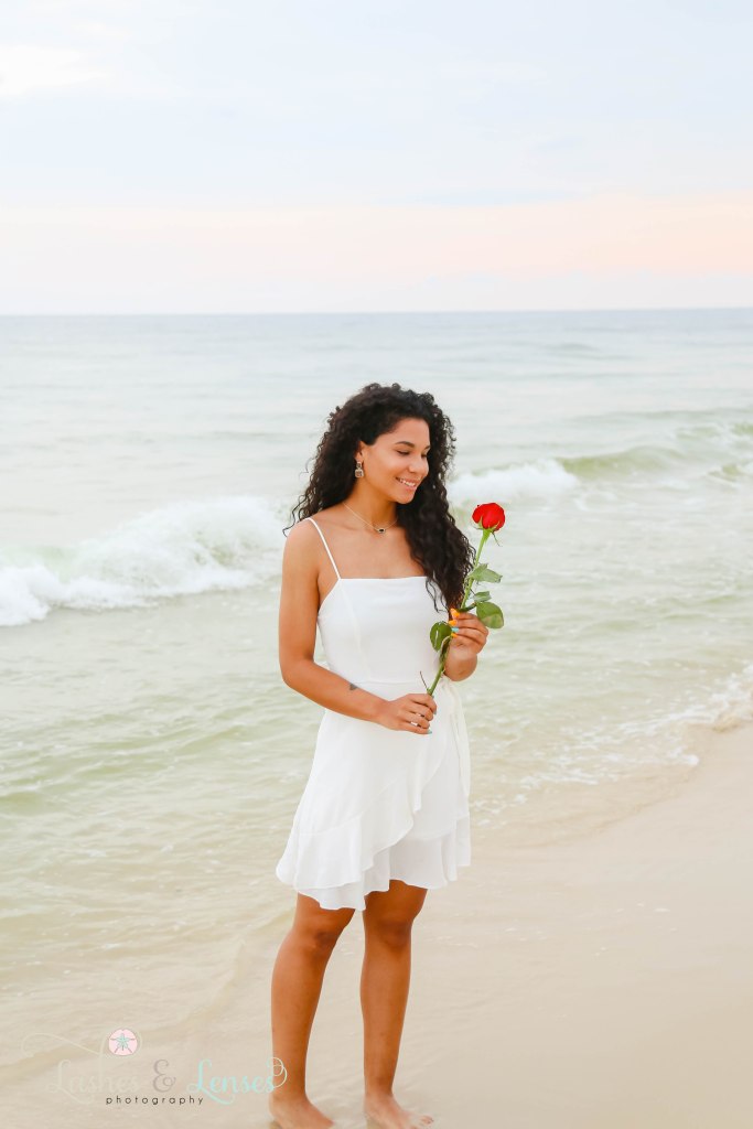 Young adult women holding a long stem red rose with the waves crashing behind her at Johnsons Beach in Perdido Key Florida