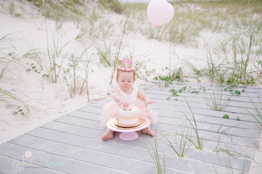 One year old little redheaded girl with a birthday crown on her head doing a cake smash, sitting on the boardwalk at Johnson's Beach in Perdido Key, Florida