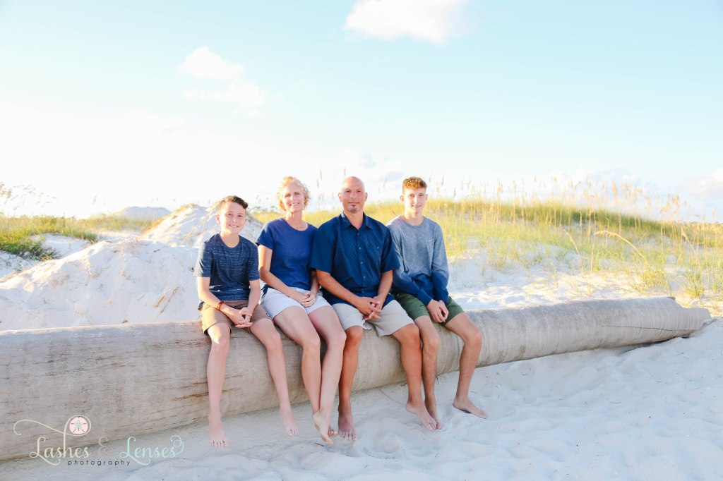 Mom, Dad and their two sons sitting on a washed up palm tree on the beach at Johnsons Beach in Perdido Key Florida