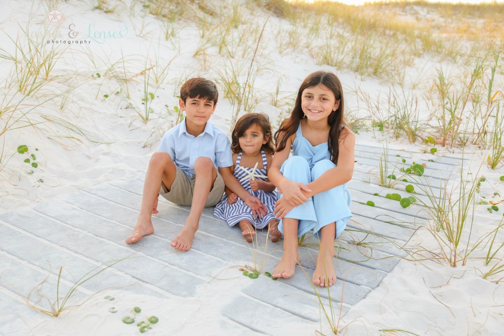 Two sisters and brother sitting on a washed up boardwalk with the sand dunes behind them at Johnsons Beach in Perdido Key, Fl