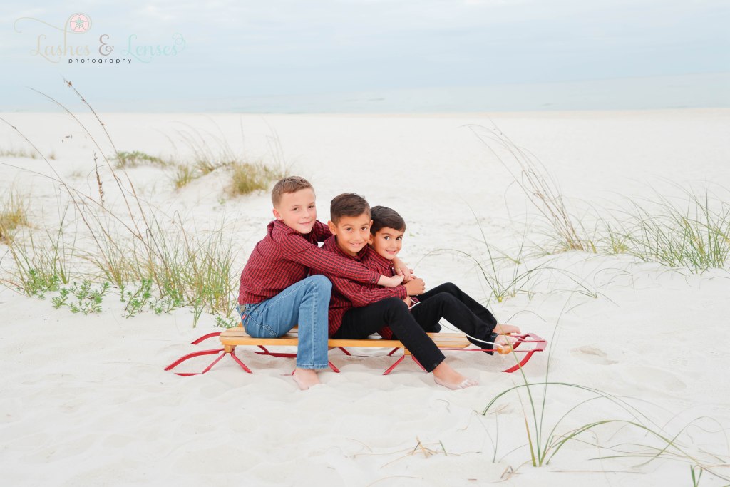 Three boy cousins all sitting on a radio flyer sled in the sand with the water behind them at Johnsons Beach in Perdido Key, Fl