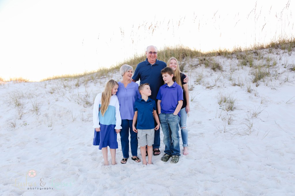 Grandparents with their four grandchildren, they are all looking at each other and smiling with the sand dunes behind them at Johnsons Beach in Perdido Key, Fl