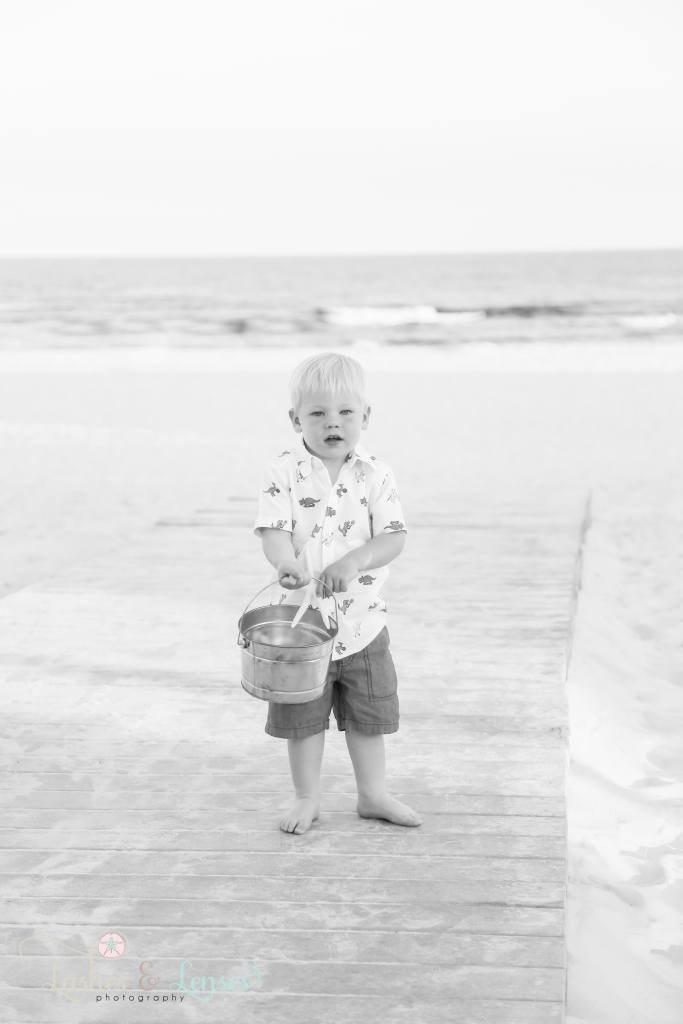 Black and White photo of little boy holding a bucket with the water behind him and standing on a boardwalkat Johnsons Beach in Perdido Key, Fl