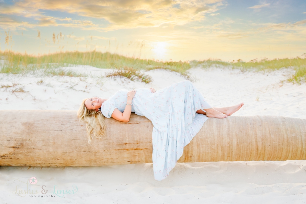 Pregnant mom laying on a washed up palm tree and holding her belly with the sunset behind her at Johnsons Beach in Perdido Key, Fl