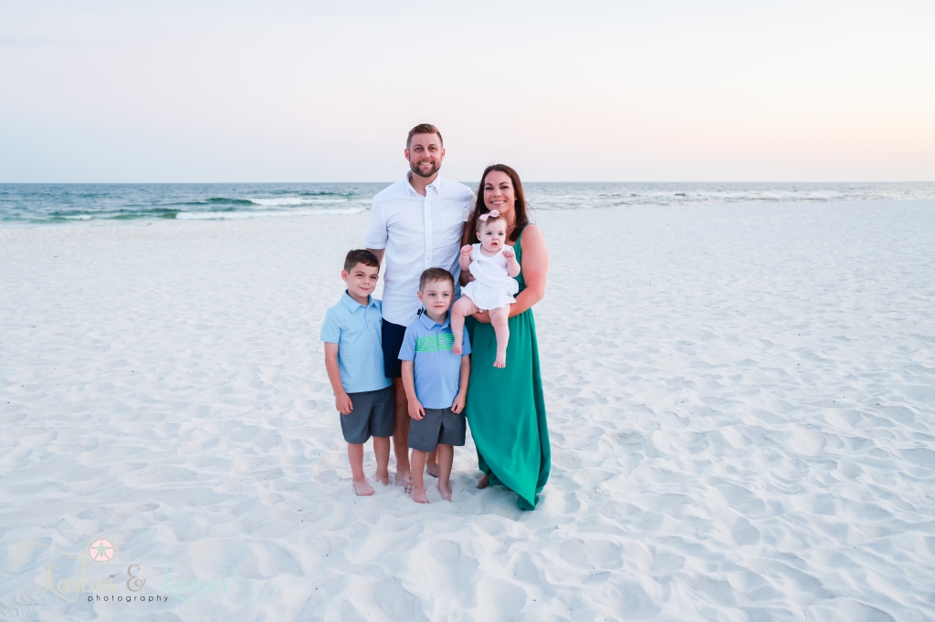 Mom and Dad standing on the beach with the water behind them, their two young boys are standing with them and mom is holding baby girl at Johnsons Beach in Perdido Key, Florida