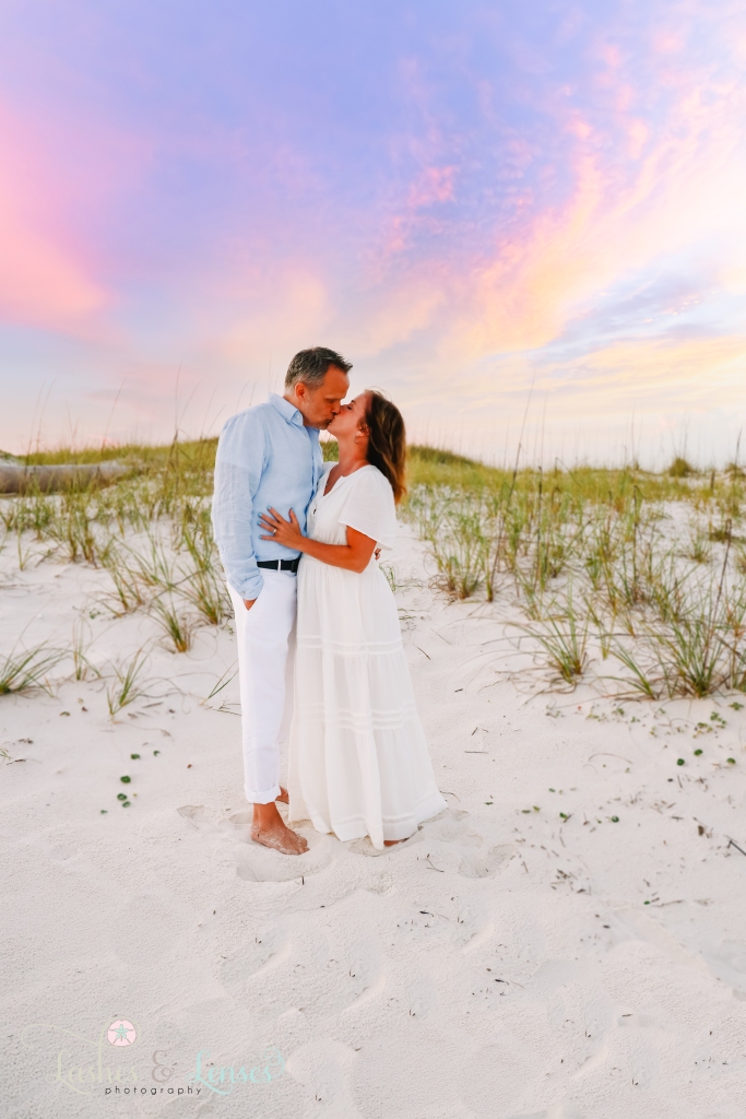 Husband and wife kissing with the sunset and sand dunes behind them at Johnsons Beach in Perdido Key, Florida