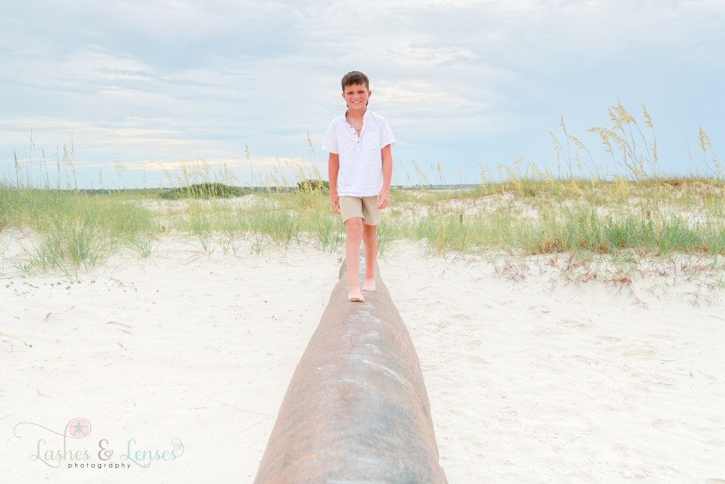 Boy walking on a washed up palm tree trunk with sand dunes and sea oats behind him at Johnsons Beach in Perdido Key, Florida