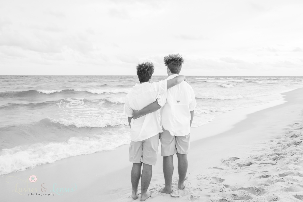 Black and white photo of two teen brothers with their arms around each other and with their backs to the camera. They are looking out at the water at Johnson's Beach in Perdido Key. Florida