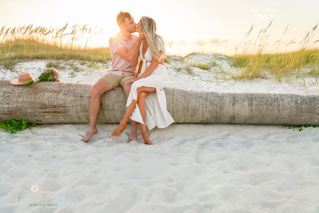 Couple sitting on a washed up palm tree, they are kissing with the sun flare behind them at Johnsons Beach in Perdido Key, Florida