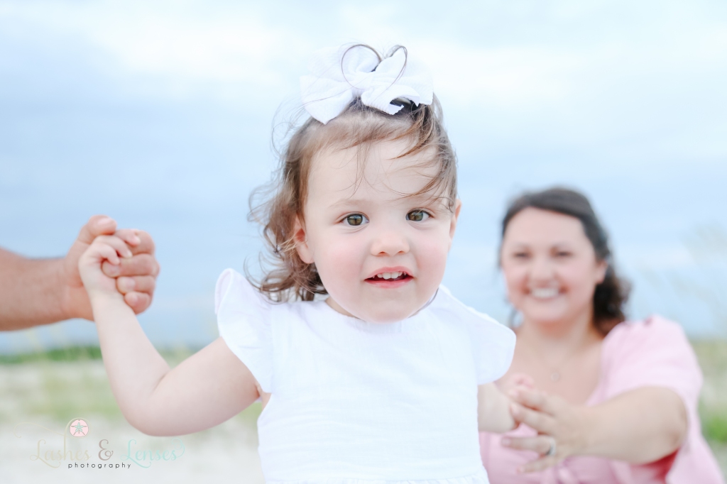 Close up photo of toddler girl smiling and looking into the camera with mom blurred out behind her at Johnson's Beach in Perdido Key, Florida