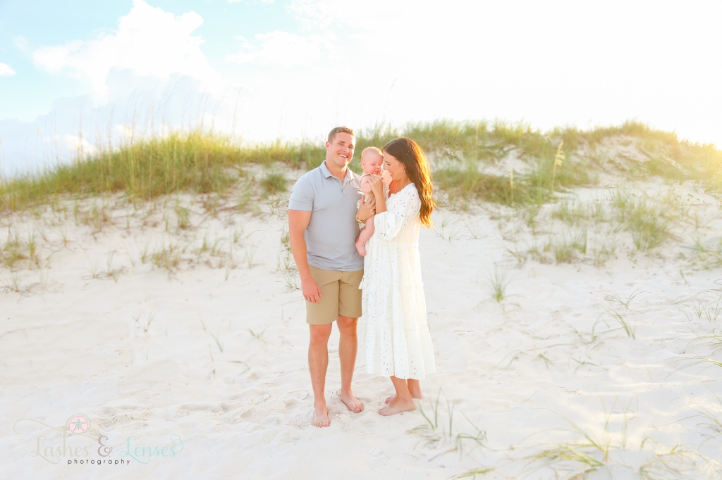 Husband and wife standing on the beach and holding their baby son, there are sand dunes and sea oats behind them at Johnsons Beach in Perdido Key, Florida