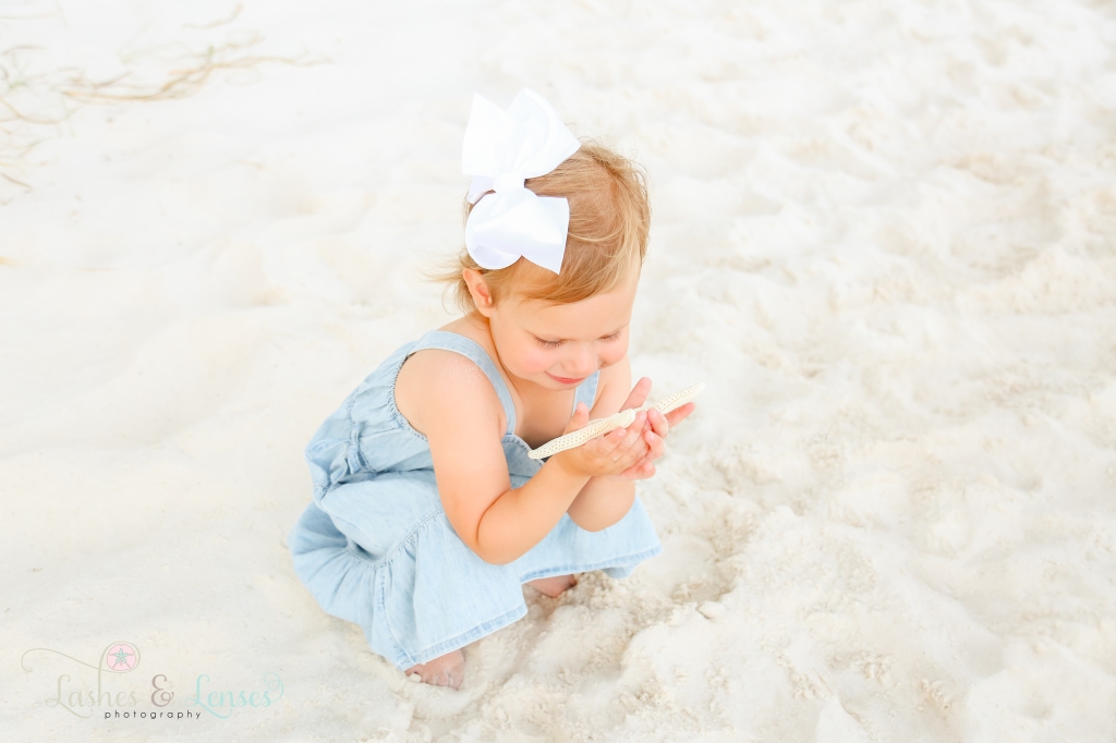 Toddler girl sitting in the sand and looking down at the starfish in her hands at Johnsons Beach in Perdido Key, Florida