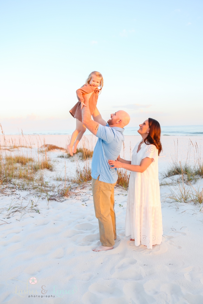 Dad holding little girl up in the air with mom standing behind them at Johnsons Beach in Perdido Key, FL