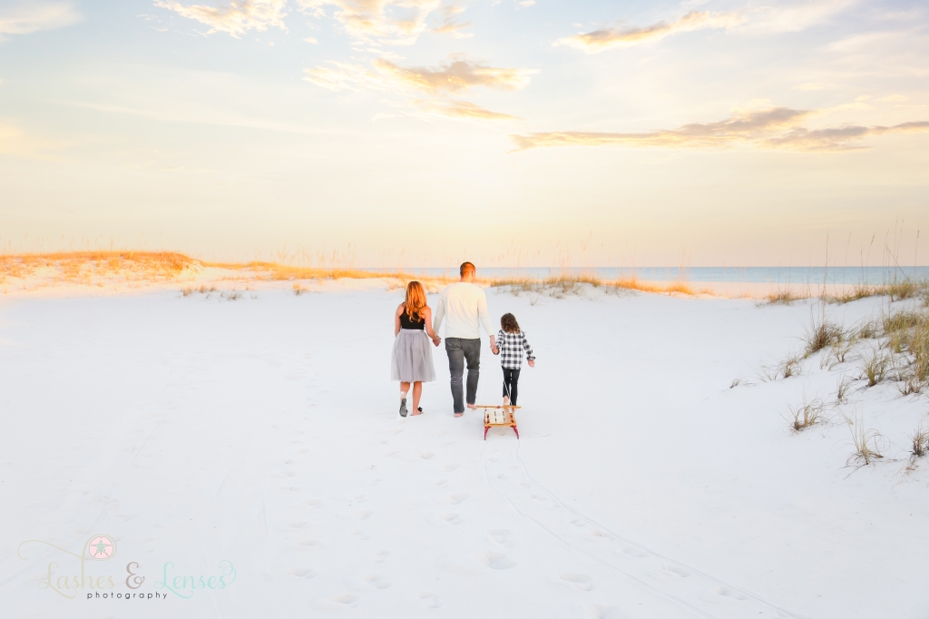 Mom and Dad holding hands with their son and walk on the beach and pulling a wood radio flyer sled at Johnsons Beach in Perdido Key, FL